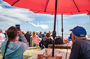 People watching planes in the sky under a large sun umbrella